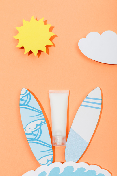 Top view of paper cut sun, cloud, sea wave and surfboards with tube of sunscreen on orange  - Photo, Image