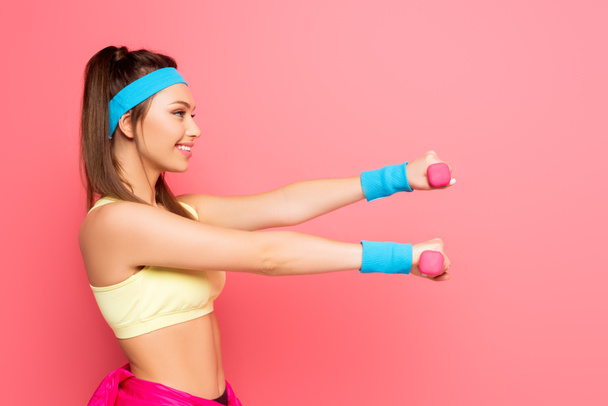 boční pohled na usmívající se mladé sportovkyně školení s činky na růžovém pozadí - Fotografie, Obrázek