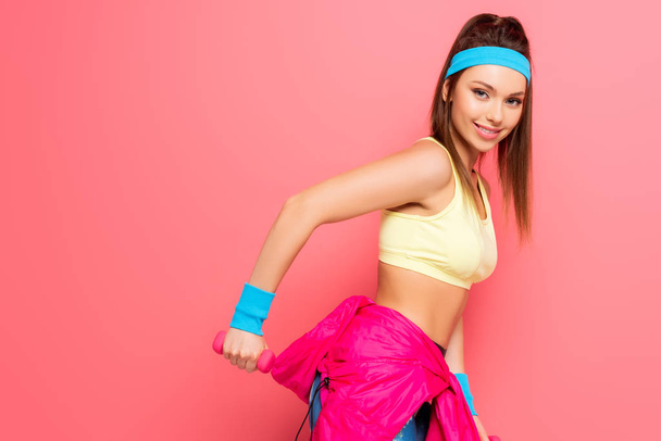 beautiful young sportswoman training with dumbbell while smiling at camera on pink background - Photo, Image