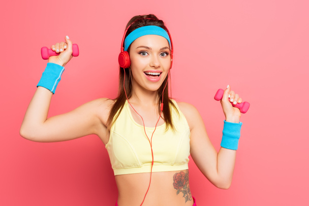 veselá sportovkyně ve sluchátkách školení s činky na růžovém pozadí - Fotografie, Obrázek