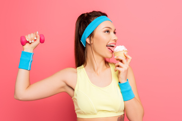 щаслива спортсменка тримає гантелі і їсть смачний кекс на рожевому фоні
 - Фото, зображення