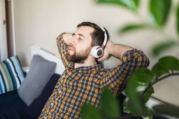 Man luisterend naar muziek in koptelefoon op de bank thuis, met zijn ogen dicht en handen achter het hoofd. - Foto, afbeelding