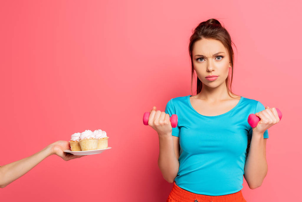 confident sportswoman exercising with dumbbells near female hand with plate of delicious cupcakes on pink background - Foto, Bild