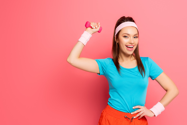 happy sportswoman exercising with dumbbell while holding hand on hip on pink background - Fotoğraf, Görsel