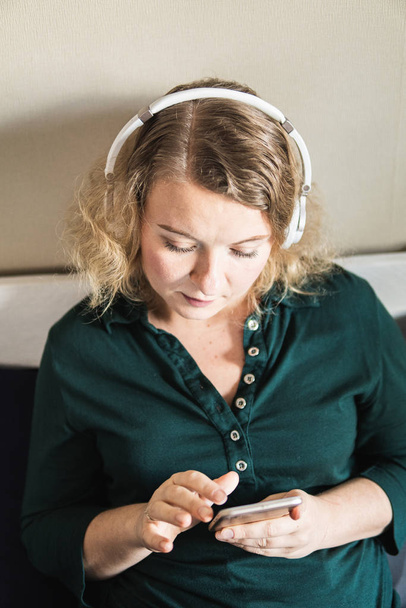 Genç sarışın kadın evdeki koltukta kulaklıkla müzik dinliyor, cep telefonu ekranına bakıyor. Dikey yönelim - Fotoğraf, Görsel