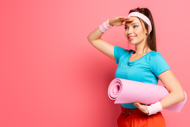 smiling sportswoman looking away while holding fitness mat on pink background - Photo, Image