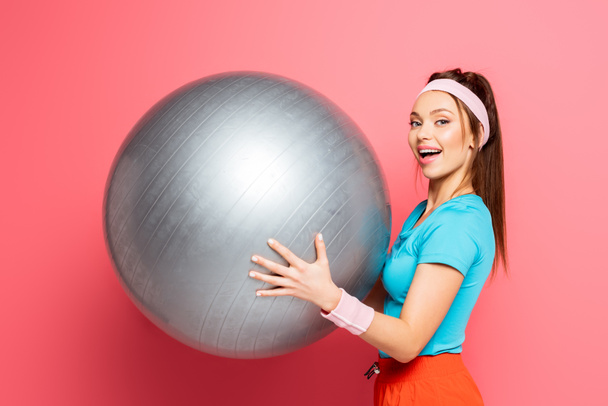 ピンクを背景にカメラで笑顔しながらシルバーフィットネスボールを持っている陽気なスポーツ女性 - 写真・画像