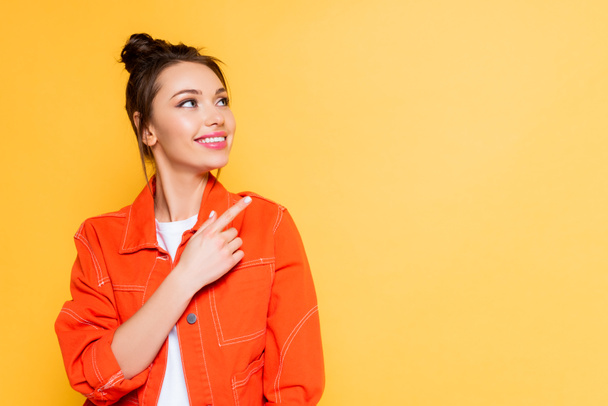 attractive smiling woman looking away and pointing with finger isolated on yellow - Фото, изображение