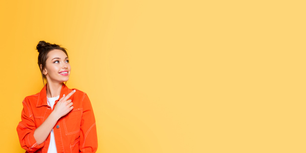 plano panorámico de mujer sonriente atractiva mirando hacia otro lado y señalando con el dedo aislado en amarillo
 - Foto, Imagen