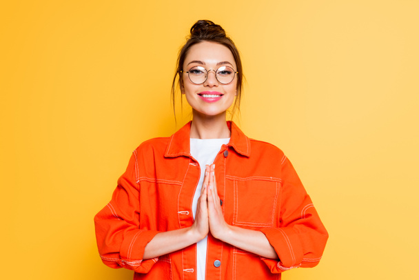 smiling student in eyeglasses standing with praying hands isolated on yellow - Photo, Image
