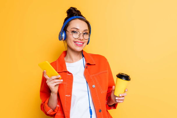 smiling student in headphones holding smartphone and coffee to go on yellow isolated on yellow - Photo, Image