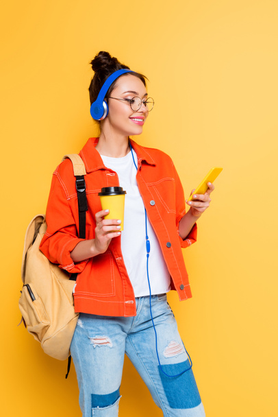 estudante alegre em fones de ouvido usando smartphone enquanto segurando café para ir no fundo amarelo
 - Foto, Imagem