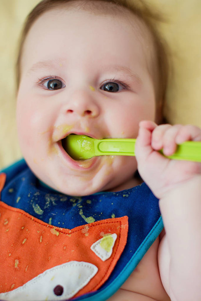 Pikkupoika syö ensimmäisen ruokansa vihreästä lusikasta. Vauva söpö ruokalappu syödä vihannes sose
 - Valokuva, kuva