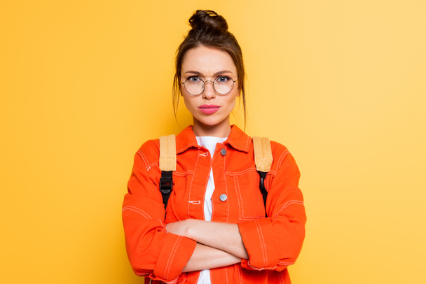 displeased student in eyeglasses standing with crossed arms while looking at camera on yellow background  - Photo, Image