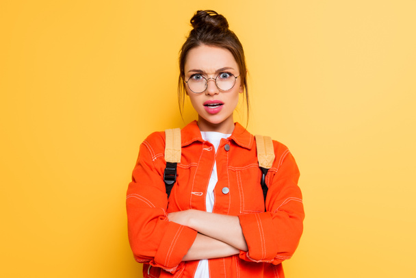 dissatisfied student in eyeglasses standing with crossed arms while looking at camera on yellow background  - Фото, изображение