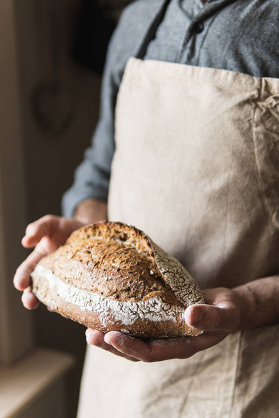 Man in rustic apron holding a loaf of bread in his hands. Close up on the product. - Photo, Image