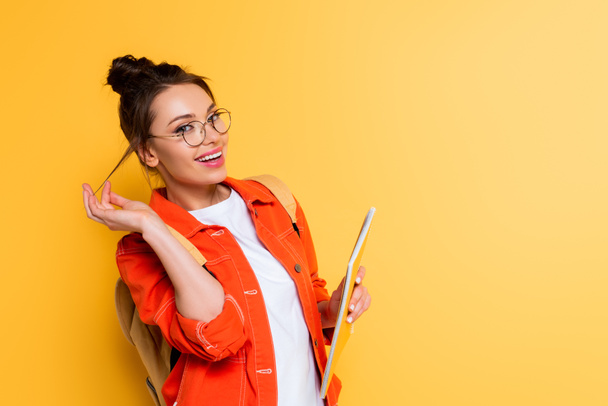 flirty student touching hair while holding notebook and smiling at camera on yellow background - Foto, afbeelding