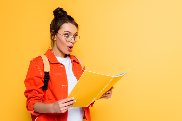 shocked student in glasses looking at copy book isolated on yellow - Photo, image