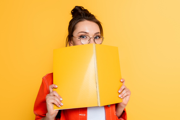young student in glasses obscuring face with copy book while looking at camera isolated on yellow - Foto, Bild