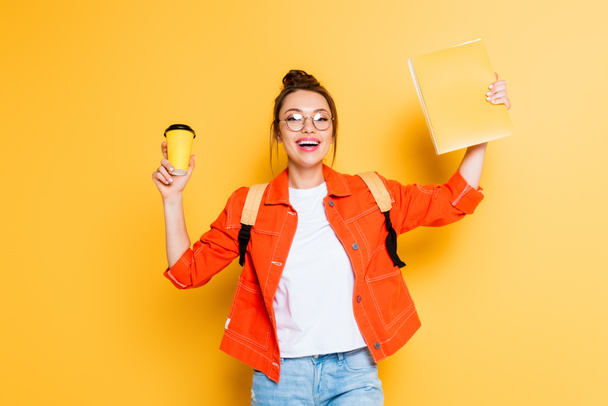 excited student holding coffee to go and copy book in raised hands while smiling at camera on yellow background - Valokuva, kuva