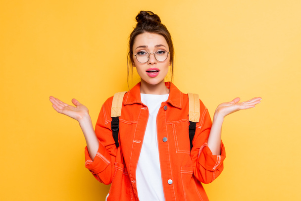 confused student in eyeglasses showing shrug gesture while looking at camera  on yellow background - Foto, Imagem