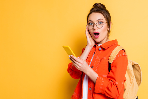 Schockierter Student mit Brille berührt Gesicht beim Chatten auf Smartphone auf gelbem Hintergrund - Foto, Bild