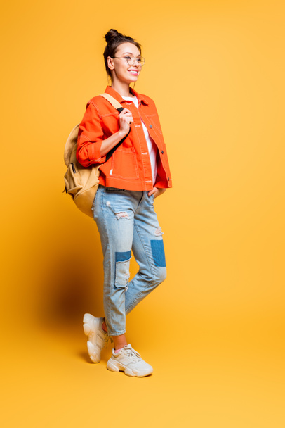 full length view of smiling student with backpack looking away while holding hand in pocket on yellow background - Photo, Image