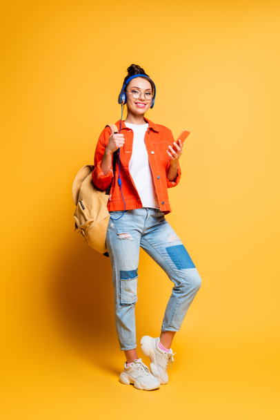 attractive smiling student in headphones holding backpack and smartphone while looking at camera on yellow background - Фото, зображення