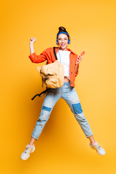 cheerful student in headset and glasses jumping while holding smartphone on yellow background - Valokuva, kuva