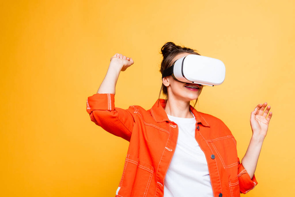 smiling girl gesturing while using vr headset isolated on yellow - Foto, Bild
