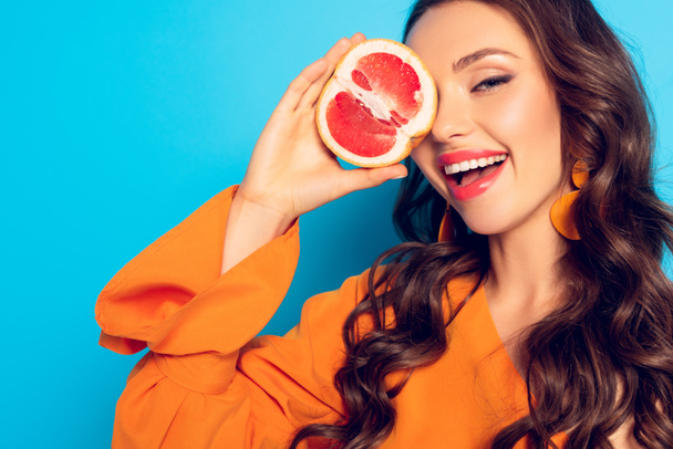 cheerful girl covering eye with half of ripe pineapple on blue background - Fotografie, Obrázek