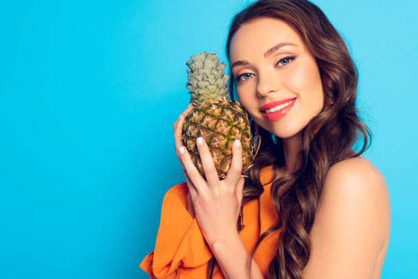 mooi meisje glimlachen op camera terwijl het houden van rijpe ananas op blauwe achtergrond - Foto, afbeelding