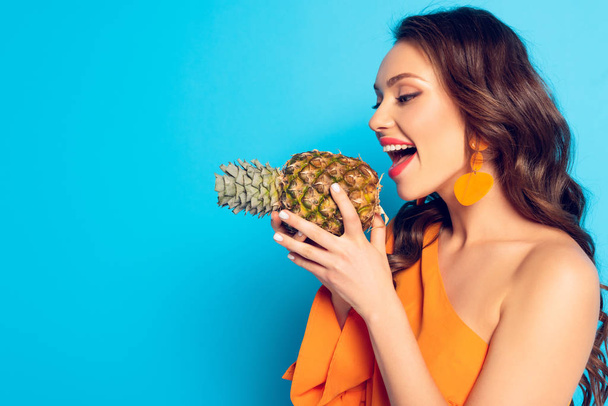 Neşeli kız, ağzı açık, mavi arka planda olgun ananas tutuyor. - Fotoğraf, Görsel