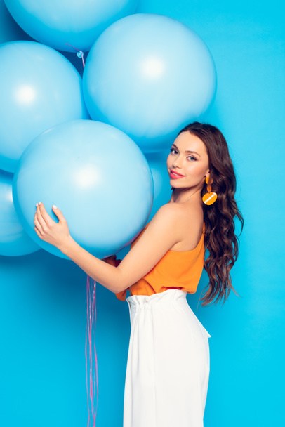 beautiful stylish girl smiling at camera while holding big festive balloons on blue background - Fotoğraf, Görsel