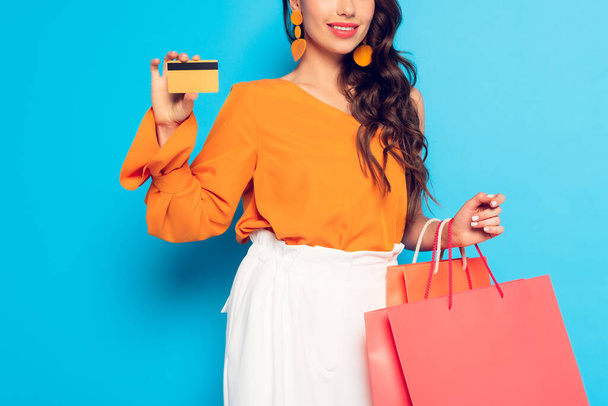 vista recortada de una chica de moda sosteniendo bolsas de compras y mostrando la tarjeta de crédito sobre fondo azul
 - Foto, imagen