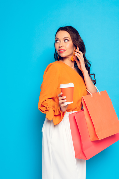 attractive elegant girl holding coffee to go and shopping bags while talking on smartphone away on blue background - Fotografie, Obrázek