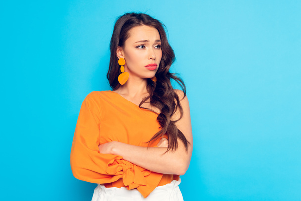upset, fashionable girl looking away while standing with crossed arms on blue background - Foto, Imagem