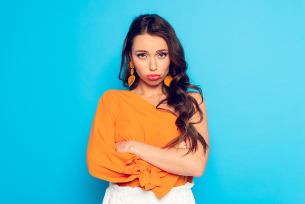 offended, pouting girl looking at camera while standing with crossed arms on blue background - Photo, image