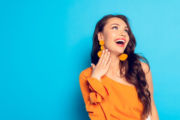 trendy, excited girl looking away while holding hand near neck on blue background - Фото, изображение