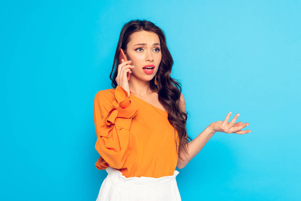 attractive, offended girl talking on smartphone while standing with open arm isolated on blue - Photo, Image