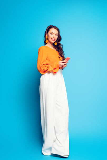 full length view of beautiful, happy woman in stylish clothes using smartphone and smiling at camera on blue background - Fotografie, Obrázek