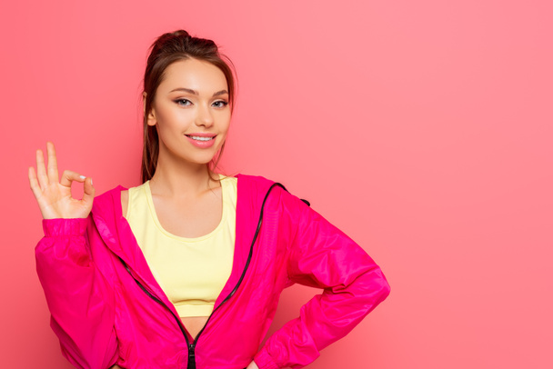 beautiful, smiling sportswoman showing okay gesture while looking at camera on pink background - Photo, image