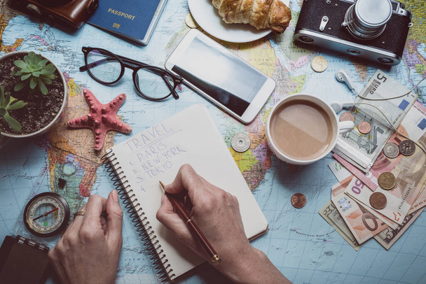 Plan of trip. background - what to take for a trip. Women hands and notepad for writing ideas, map, retro camera, money, coins, croissant, coffee, pen, compass, sunglasses, smartphon - Photo, Image