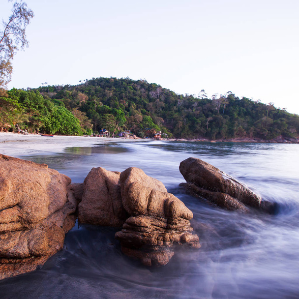 Brązowe kamienie na tropikalnej piaszczystej plaży o zmierzchu, miękki zachód słońca świeci na kształt dużych kamieni i delikatnie fal, scena letnia na wyspie Koh Chang, Ranong, Tajlandia. Długotrwałe narażenie. - Zdjęcie, obraz