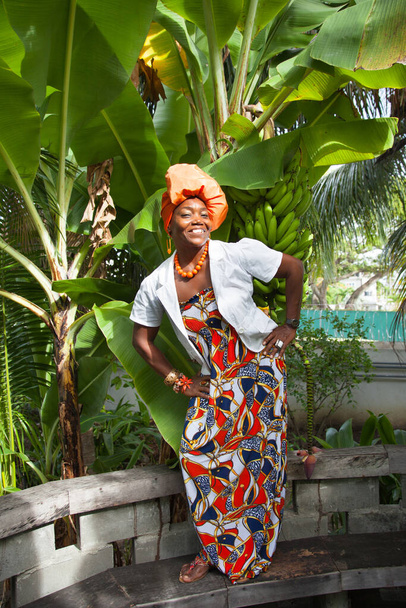 The vertical full body of a joyful African American woman wearing a bright colorful national dress poses in the garden against the background of banana trees. Traditions fashion costumes. - 写真・画像