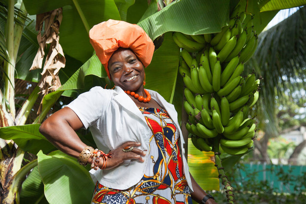 The vertical full body of a joyful African American woman wearing a bright colorful national dress poses in the garden against the background of banana trees. Traditions fashion costumes. - Foto, imagen