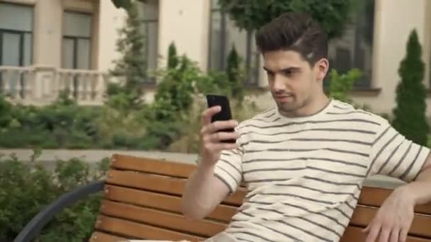 Веселий чоловік дивиться на камеру свого мобільного телефону, сидячи на лавці на міській вулиці
 - Кадри, відео