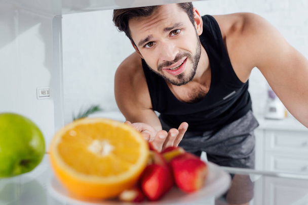 Selektiver Fokus eines verwirrten Mannes, der in die Kamera blickt und in der Nähe von Früchten im Kühlschrank gestikuliert  - Foto, Bild