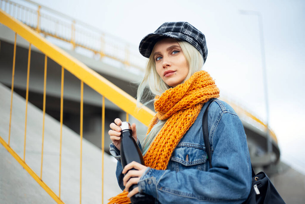 Portrait de jeune adolescente blonde, veste en denim bleu, avec écharpe orange et capuchon à carreaux, tenant une bouteille d'eau thermo eco en acier noir
. - Photo, image