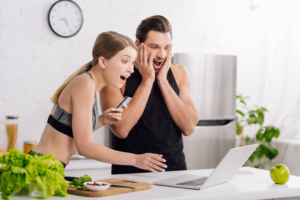 surprised man and woman looking at laptop while online shopping in kitchen  - Photo, Image